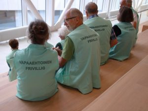 OLKAn vapaaehtoisia Siltasairaalassa. Vapaaehtoiset istuvat yhdessä puisilla portailla juttelemassa, ja heillä on yllään vaaleanvihreät liivit, joiden selässä lukee Vapaaehtoinen - Frivillig.