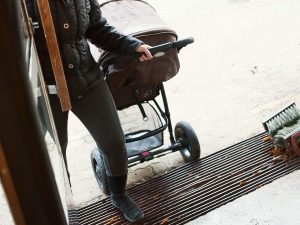 Henkilö vetää lastenvaunuja talon rapusta sisään, pitäen toisella kädellään ovenkahvaa auki.