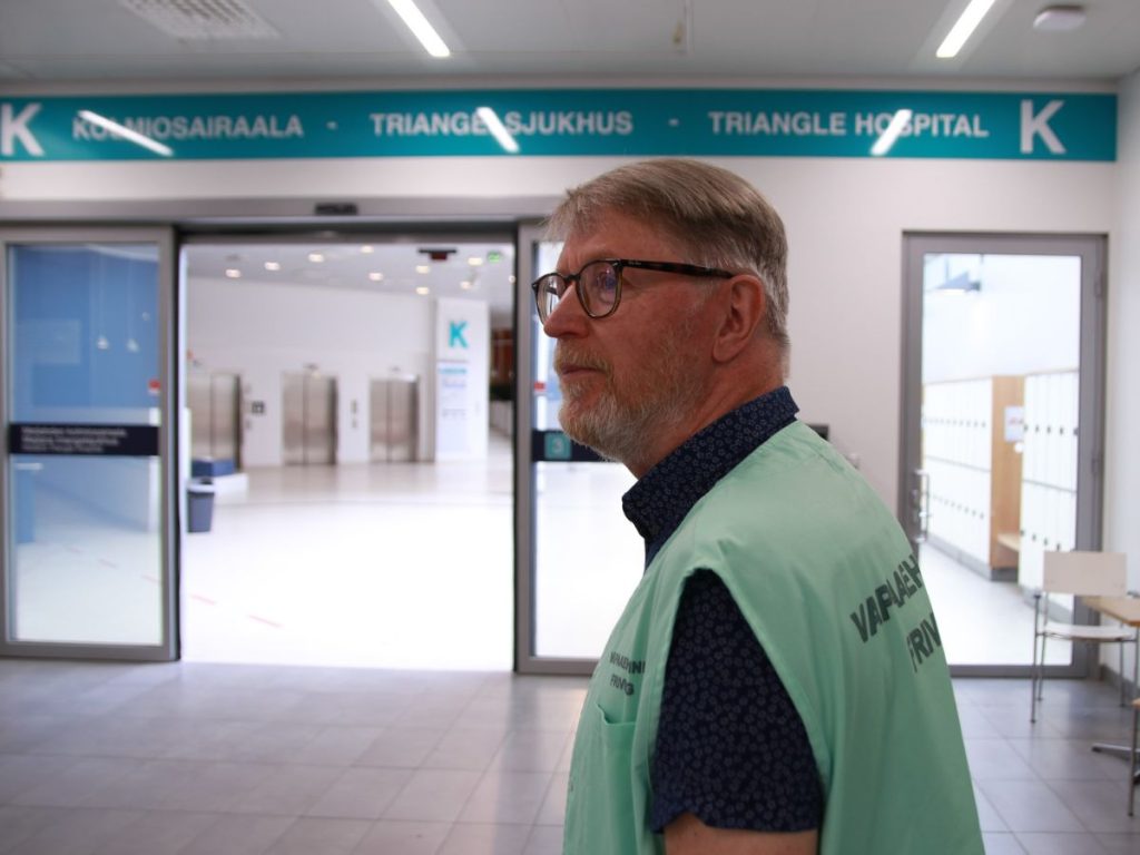 OLKAn vapaaehtoinen Meilahden sairaalan aulassa. Vapaaehtoinen on sivuttain kohti kameraa, katse pääoville suunnattuna. Hänellä on yllään vaaleanvihreä liivi, jossa lukee "vapaaehtoinen".