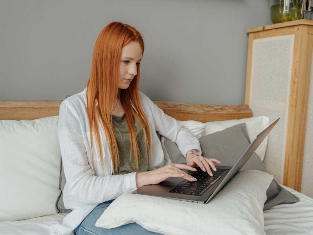 Punatukkainen nainen istuu sohvalla tietokoneen kanssa. / En rödhårig kvinna sitter i en soffa med en dator. / A red-haired woman is sitting on a sofa with a computer.