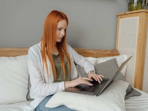Punatukkainen nainen istuu sohvalla tietokoneen kanssa. / En rödhårig kvinna sitter i en soffa med en dator. / A red-haired woman is sitting on a sofa with a computer.